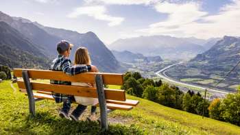 Paar auf einer Holzbank mit Aussicht auf das Rheintal und die Berge.