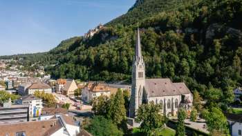 Luftaufnahme der Kathedrale St. Florin in Vaduz mit Schloss Vaduz im Hintergrund an einem sonnigen Tag.