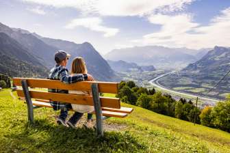 Paar sitzt auf einer Bank in Triesenberg und blickt auf das Rheintal in Liechtenstein mit beeindruckendem Alpenpanorama