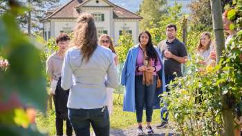 Führung in der Hofkellerei die eine Frau zeigt wie sie der Gruppe in mitten von Weinreben über den Wein und die Kultur informiert