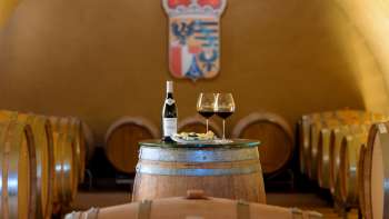 Weinfasskeller mit Flasche Rotwein und zwei Gläsern vor dem Wappen der Hofkellerei in Liechtenstein