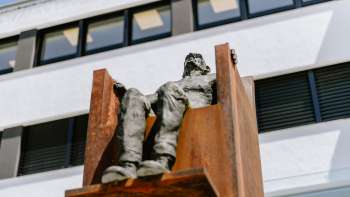 Skulptur „Hochsitz“ von Robert Indermaur: Figur sitzt erhöht auf rostfarbenem Stahlstuhl vor moderner Fassade. Skulptur „Hochsitz“ von Robert Indermaur: Figur sitzt erhöht auf rostfarbenem Stahlstuhl vor moderner Fassade.