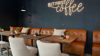 Ein modernes Café mit Ledersofas, weißen Sesseln und Holztischen. An der dunkelblauen Wand leuchtet der Schriftzug „BUT FIRST coffee“ in heller Neon-Schrift.