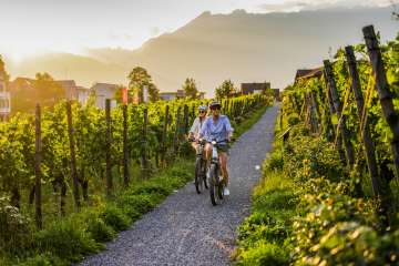 Zwei Radfahrerinnen in den Weinbergen von Liechtenstein bei Sonnenuntergang – E-Bike-Tour mit Blick auf die Alpen und Reben