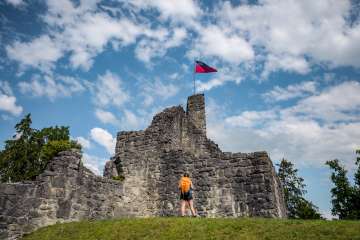 Wanderin vor der historischen Burgruine Schellenberg mit wehender Liechtenstein-Flagge unter blauem Himmel