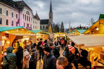 Stimmungsvolle Abendaufnahme des Weihnachtsmarkts in Vaduz 2018 mit festlich geschmückten Marktständen und Menschenmenge vor Regierungsgebäude und Kathedrale