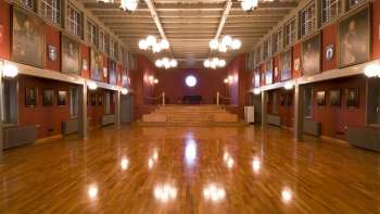 Historischer Rathaussaal in Vaduz mit glänzendem Holzfußboden, roten Wänden, Porträtgemälden und prunkvollen Kronleuchtern.