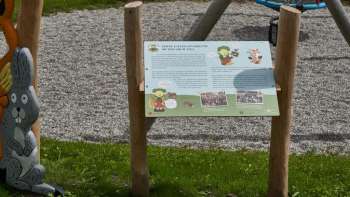 Zwei Kinder betrachten gemeinsam eine Erlebnisstation und lernen auf dem interaktiven Naturpfad „Wurz und Zirp auf Zeitreise“ über Tiere und Pflanzen in Liechtenstein.