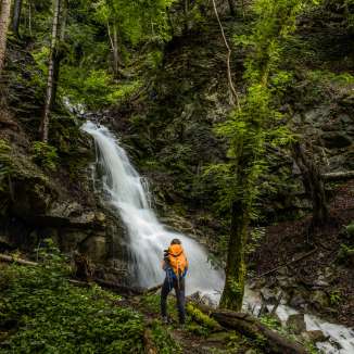 Wanderer mit Rucksack vor einem idyllischen Wasserfall bei Nendeln, eingebettet in dichten Wald und moosbedeckte Felsen.