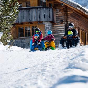 Zwei Erwachsene und drei Kinder sitzen auf Schlitten und starten gemeinsam eine Abfahrt vor einer verschneiten Berghütte in Malbun
