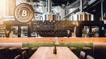 Gemütlicher Schankraum im Liechtensteiner Brauhaus mit Holztischen, Zapfanlage und Blick auf die Sudkessel